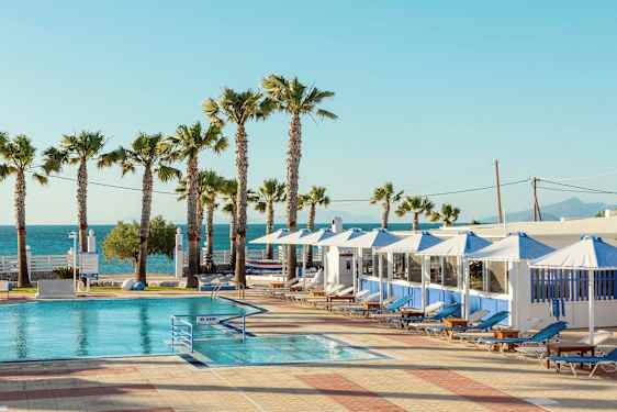 Uima-allas aurinkotuoleilla ja sateenvarjoilla kirkkaan taivaan alla. Taustalla palmuja ja meri. (Tekoälyn luoma teksti)