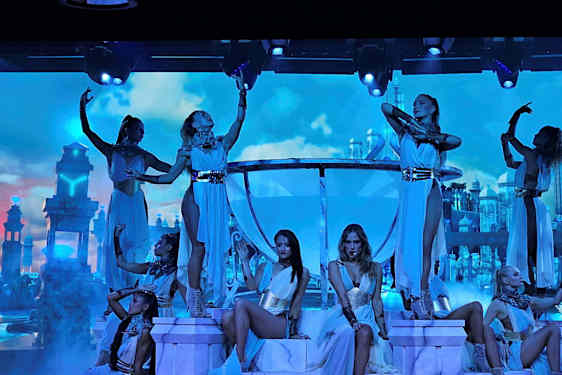 Tanssijat poseeraavat valaistulla näyttämöllä palatsimaisia rakennuksia sisältävän taustan edessä pilvien täyttämällä taivaalla. (Tekoälyn luoma teksti)