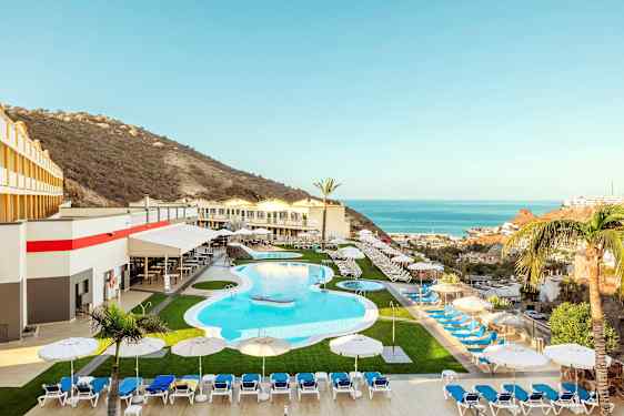 Lomahotelli, jossa on uima-allas, aurinkotuoleja ja -varjoja, ympäröitynä vuoristomaisemilla ja merinäköalalla. (Tekoälyn luoma teksti)