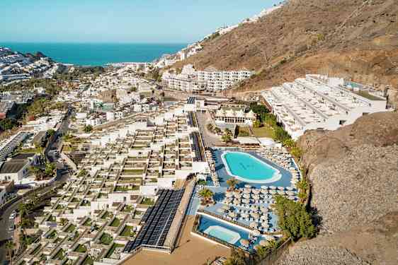 Ilmakuva hotellista, jossa on uima-altaita, aurinkotuoleja ja terasseja, sijaitsee rinteessä lähellä rannikkoa. (Tekoälyn luoma teksti)