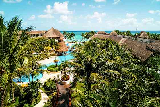 Suuri lomakeskus, jossa on uima-altaita, palmuja ja olkikattoisia rakennuksia, sijaitsee lähellä merta sinisen taivaan alla. (Tekoälyn luoma teksti)