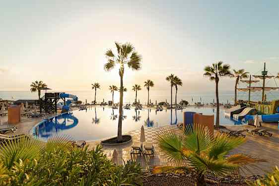 Kuva uima-altaasta, vesiliukumäistä, aurinkotuoleista ja palmuista sekä taustalla näkyvästä merestä auringonlaskun aikaan. (Tekoälyn luoma teksti)