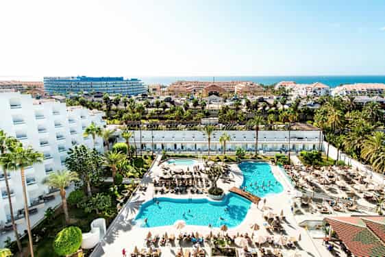 Näkymä hotellialueelle, jossa uima-altaita, aurinkotuoleja, palmuja ja rakennuksia taustalla. (Tekoälyn luoma teksti)