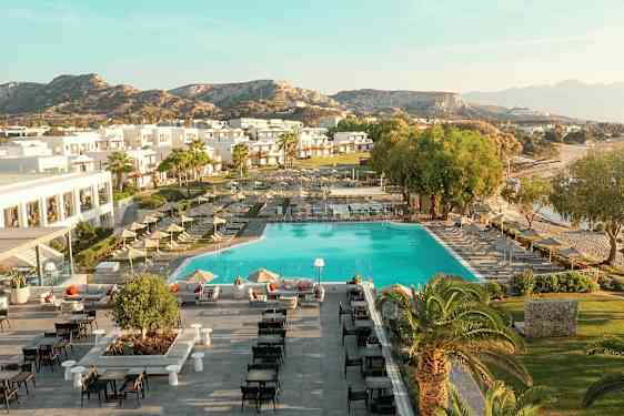 Ilmakuva lomakeskuksesta, jossa uima-allas, aurinkotuoleja, rakennuksia ja ympäröivä maisema. (Tekoälyn luoma teksti)