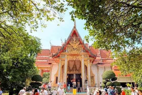 Perinteinen rakennus, jossa on koristeellinen punainen ja kultainen kattorintama, ympäröitynä vihreillä puilla ja ihmisillä. (Tekoälyn luoma teksti)