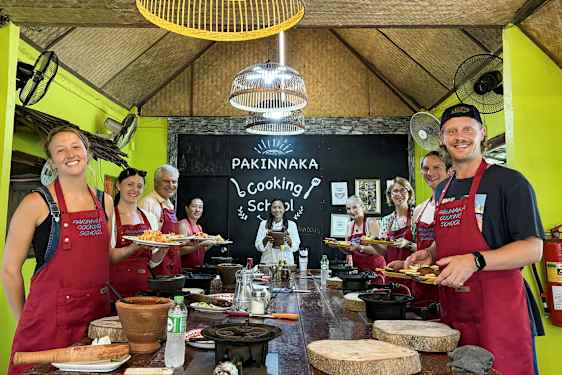 Ryhmää ihmisiä esiliinoissaan seisoo kyltin edessä, jossa lukee 'Pakinnaka Cooking School' keittiöstudiossa. (Tekoälyn luoma teksti)