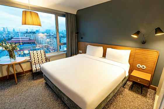 Hotellihuone, jossa suuri sänky, ikkuna ja näkymä kaupunkiin. (Tekoälyn luoma teksti)