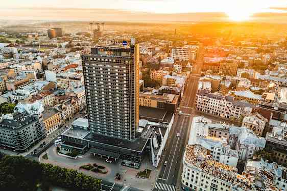 Ilmakuva kaupungista, jonka etualalla on korkea rakennus ja ympärillä muita rakennuksia, auringon laskiessa horisontissa. (Tekoälyn luoma teksti)