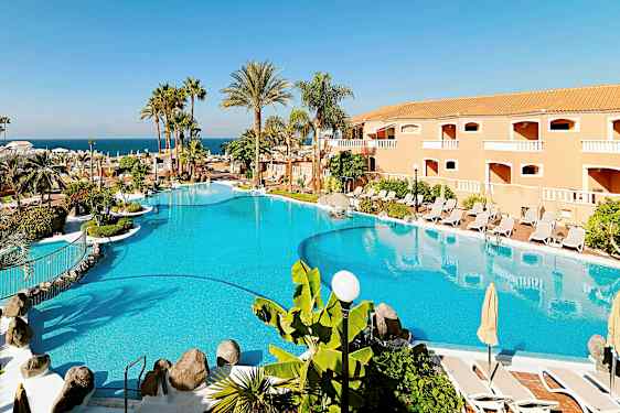 Suuri uima-allas, jota ympäröivät aurinkovarjot, aurinkotuolit, palmut ja taustalla oleva rakennus. (Tekoälyn luoma teksti)