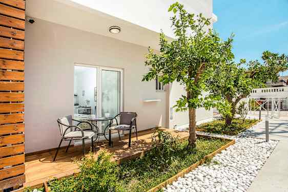 Terassi, jolla on puulattia sekä pöytä ja kaksi tuolia, ympäröitynä kasveilla ja sitruunapuulla. (Tekoälyn luoma teksti)