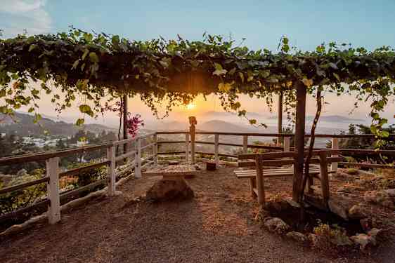 Näkötornialue, jossa puiset pergolat, jotka on peitetty lehdillä, sekä penkkejä ja kaide ympärillä. Vuoristoinen maisema auringonlaskussa taustalla. (Tekoälyn luoma teksti)