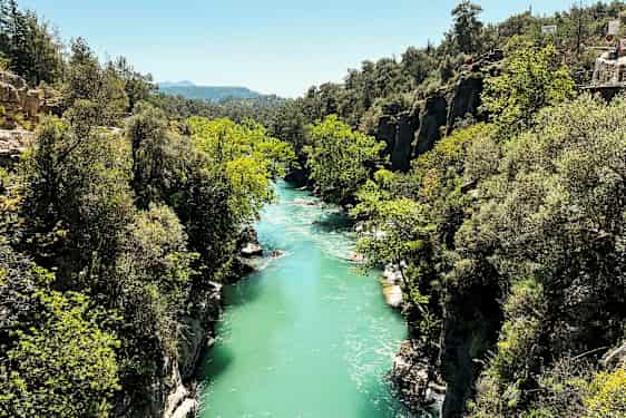 Turkoosin sininen joki virtaa vehreän rotkon läpi kallioiden ja puiden keskellä. (Tekoälyn luoma teksti)