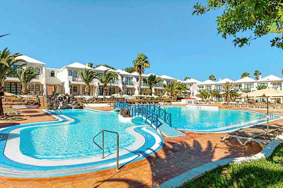 Hotelli, jossa on valkomaalattuja rakennuksia ja suuri sininen uima-allas edessä, ympärillä palmuja ja vihreitä alueita. (Tekoälyn luoma teksti)