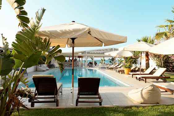 Uima-allas, jota ympäröivät aurinkotuolit, aurinkovarjot ja vehreys. Taustalla näkyy meri ja rakennus. (Tekoälyn luoma teksti)