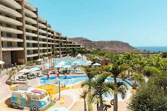 Ulkotila, jossa suuri uima-allas, aurinkotuoleja ja varjoja ympärillä sekä leikkialue, näkymä merelle ja vuoristomaisemaan. (Tekoälyn luoma teksti)