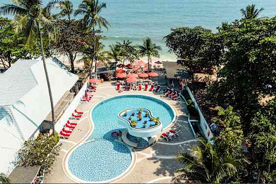 Ilmakuva uima-altaasta lomakeskuksessa, jossa on aurinkotuoleja ja varjoja. Ympärillä on trooppisia puita ja lähellä merta. (Tekoälyn luoma teksti)