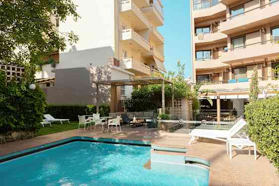 Ulkotila, jossa on uima-allas ja aurinkotuoleja rakennusten ja vihreän ympäröimänä. (Tekoälyn luoma teksti)