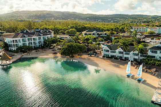 Useita hotellirakennuksia rauhallisella hiekkarannalla, jossa kirkas vesi ja näkymä vehreille mäille. (Tekoälyn luoma teksti)