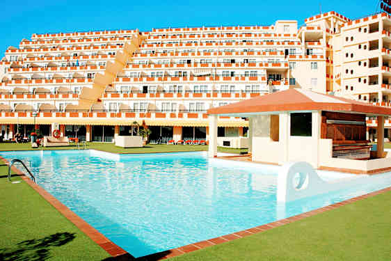 Suuri uima-allas, jota ympäröivät vihreät alueet, ja taustalla usean kerroksen hotelli. (Tekoälyn luoma teksti)