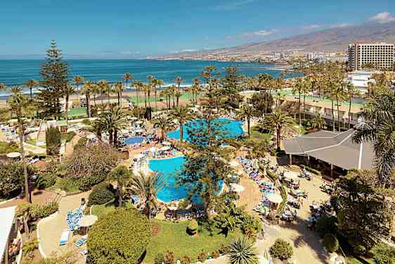 Näkymä lomakeskukseen, jossa on uima-altaita, palmuja ja ihmisiä auringonotto- ja uintipuuhissa lähellä merta. (Tekoälyn luoma teksti)