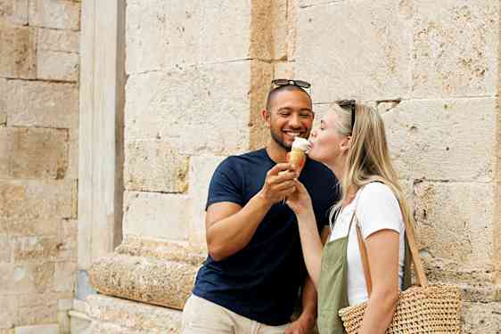 Kaksi henkilöä seisoo kivirakennuksen äärellä, joista yhdellä on olkilaukku. (Tekoälyn luoma teksti)