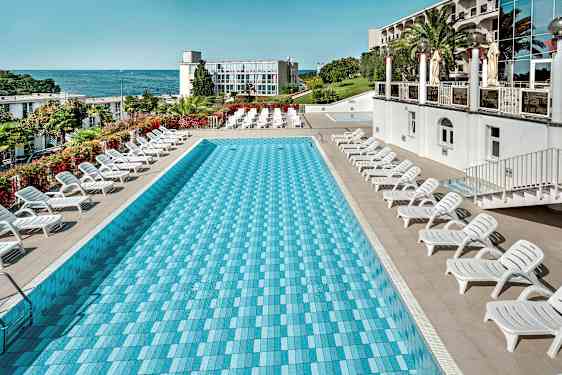 Suorakulmainen uima-allas, jonka ympärillä on aurinkotuoleja, ja taustalla meri ja rakennus. (Tekoälyn luoma teksti)