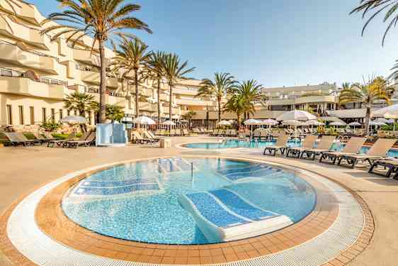 Hotellin uima-allasalue, jossa aurinkotuoleja ja aurinkovarjoja altaan ympärillä, palmupuiden ja parvekkeiden ympäröimänä. (Tekoälyn luoma teksti)