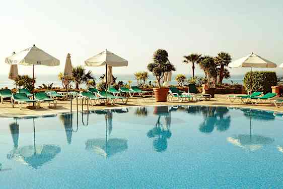 Uima-allas, jota ympäröivät aurinkotuolit ja aurinkovarjot, näkymä merelle ja ympäröiville kasveille. (Tekoälyn luoma teksti)