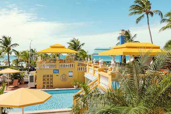 Trooppinen ympäristö, jossa uima-allas ja keltaisia aurinkovarjoja palmujen ympäröimänä, taustalla näkyy meri. (Tekoälyn luoma teksti)