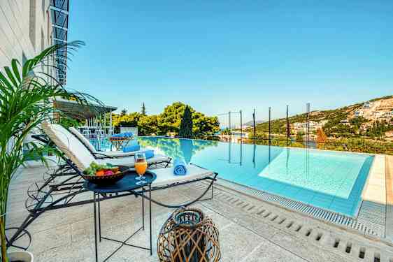 Näkymä infinity-uima-altaan suuntaan, jossa on aurinkotuoleja, pöytiä hedelmien ja juomien kanssa vieressä sekä vihreä ympäristö ja taivas taustalla. (Tekoälyn luoma teksti)