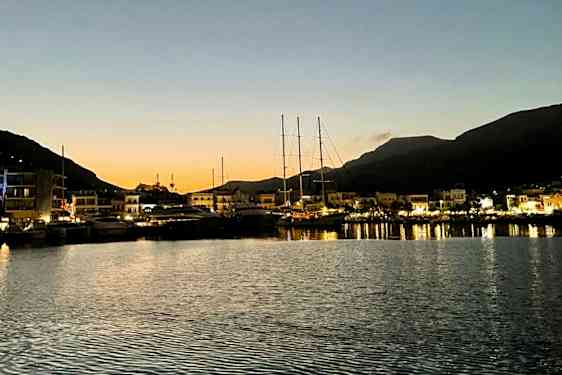 Satama hämärässä, veneitä kiinnitettynä laiturissa ja valaistuja rakennuksia taustalla. Taustalla näkyy vuoria. (Tekoälyn luoma teksti)