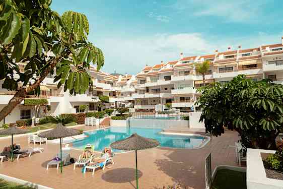 Ulkotila, jossa on uima-allas, aurinkotuoleja ja monikerroksinen hotelli. Useita aurinkovarjoja ja vihreitä kasveja ympäröivät aluetta. (Tekoälyn luoma teksti)