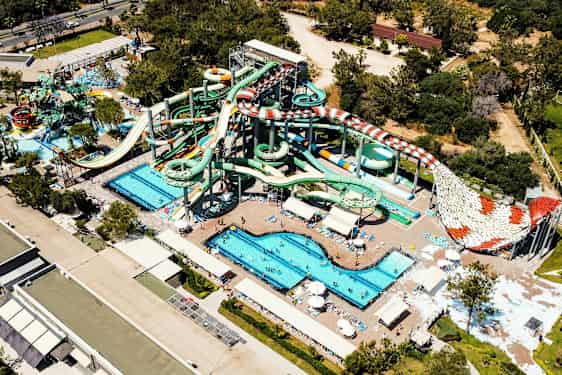 Vesipuisto, jossa on useita suuria vesiliukumäkiä, altaita ja aurinkotuoleja, ympäröitynä vihreillä puilla ja rakennuksilla. (Tekoälyn luoma teksti)