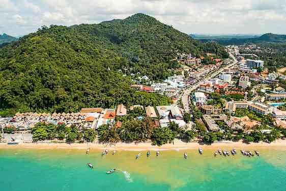 Ilmakuva rannasta, jossa veneitä vedessä, ja taustalla vehreitä kukkuloita ja kaupunkia. (Tekoälyn luoma teksti)