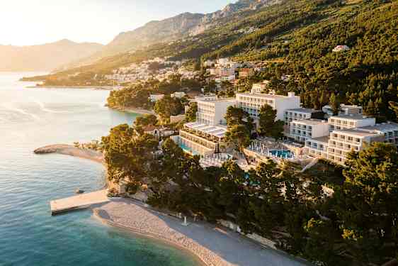 Ilmakuva hotellista rannikon ja rannan äärellä, ympärillä puita ja taustalla vuoria. (Tekoälyn luoma teksti)