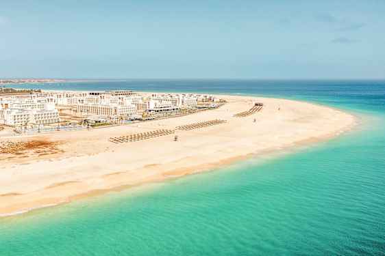 Ilmakuva valkoisesta hiekkarannasta, jossa on kirkasta turkoosia vettä ja valkoisia rakennuksia taustalla. (Tekoälyn luoma teksti)