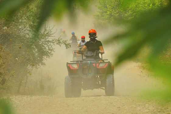 Henkilöt ajavat mönkijöillä pölyisellä soratiellä, jota ympäröi kasvillisuus. (Tekoälyn luoma teksti)