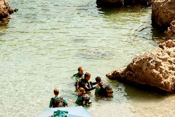 Ryhmä ihmisiä märkäpuvuissa ja sukellusvarusteissa seisoo kirkkaassa vedessä kallioiden vieressä. (Tekoälyn luoma teksti)