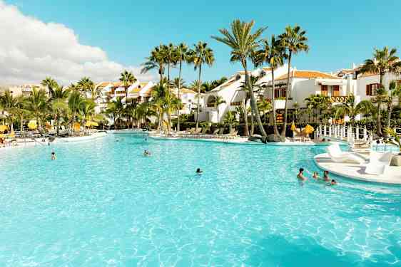 Suuri uima-allas ympäröitynä palmuilla ja valkoisilla rakennuksilla. (Tekoälyn luoma teksti)