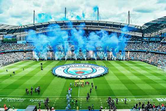 Jalkapallostadion, jossa on yleisöä ja suuri Manchester City -logolla varustettu banderolli kentällä. (Tekoälyn luoma teksti)
