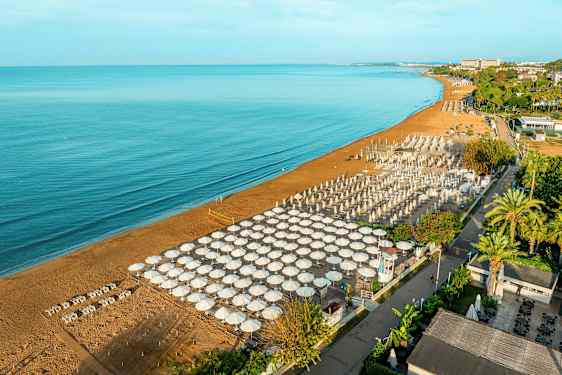 Kuvassa on hiekkaranta meren äärellä, jossa on järjestetty rivistö aurinkovarjoja ja rantatuoleja. Taustalla näkyy vihreitä puita ja rakennuksia. (Tekoälyn luoma teksti)