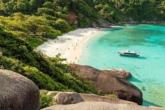 Hiekkaranta ihmisten kanssa, vehreiden puiden ympäröimä ja kirkkaan turkoosin veden reunustama. Vene on lähellä rantaa. (Tekoälyn luoma teksti)