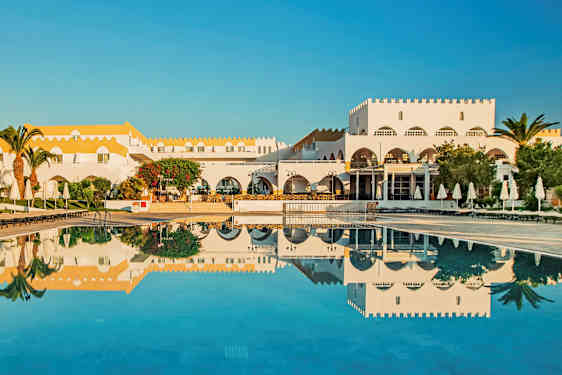 Valkoinen rakennus, jossa keltainen katto ja uima-allas, ympärillä aurinkotuoleja ja varjoja. (Tekoälyn luoma teksti)