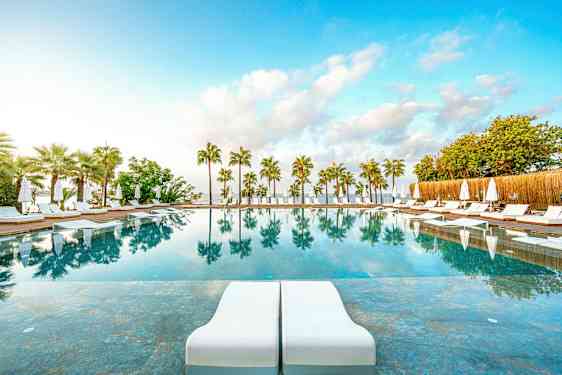 Ulkouima-allas aurinkotuoleilla ja -varjoilla, ympäröitynä palmupuilla, ja veden heijastukset näkyvät. (Tekoälyn luoma teksti)