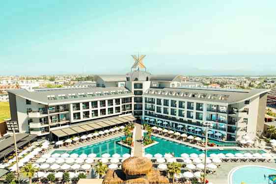 Hotellikompleksi, jossa on ulkouima-allas ja aurinkotuoleja, ympäröitynä viheralueilla ja taustalla kaupunkinäkymä. (Tekoälyn luoma teksti)