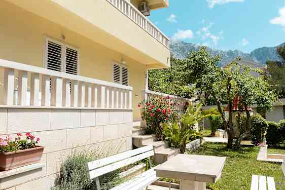 Kuva talon julkisivusta parvekkeineen ja puutarhasta etualalla, mukana penkkejä ja kukkia. (Tekoälyn luoma teksti)