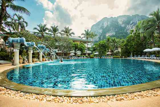 Uima-allas, jossa kirkas sininen vesi. Altaan ympärillä on palmuja ja aurinkotuoleja. Taustalla näkyy kallio ja osittain pilvinen taivas. (Tekoälyn luoma teksti)