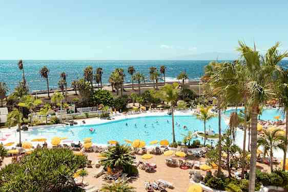 Uima-allasalue, jossa aurinkotuoleja, aurinkovarjoja ja ihmisiä, taustalla meri. (Tekoälyn luoma teksti)