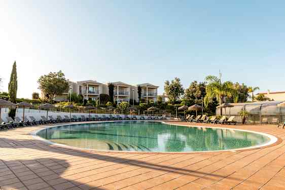 Ulkouima-allas, jossa aurinkotuoleja ja aurinkovarjoja, ympärillä vehreyttä ja rakennuksia. (Tekoälyn luoma teksti)