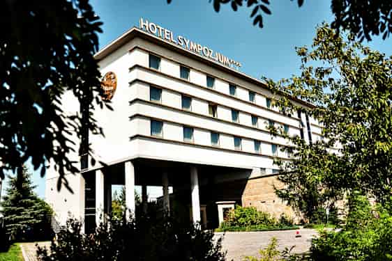 Monikerroksinen rakennus, jonka kyljessä lukee 'Hotel Sympozjum', ympäröitynä puilla ja kasvillisuudella. (Tekoälyn luoma teksti)